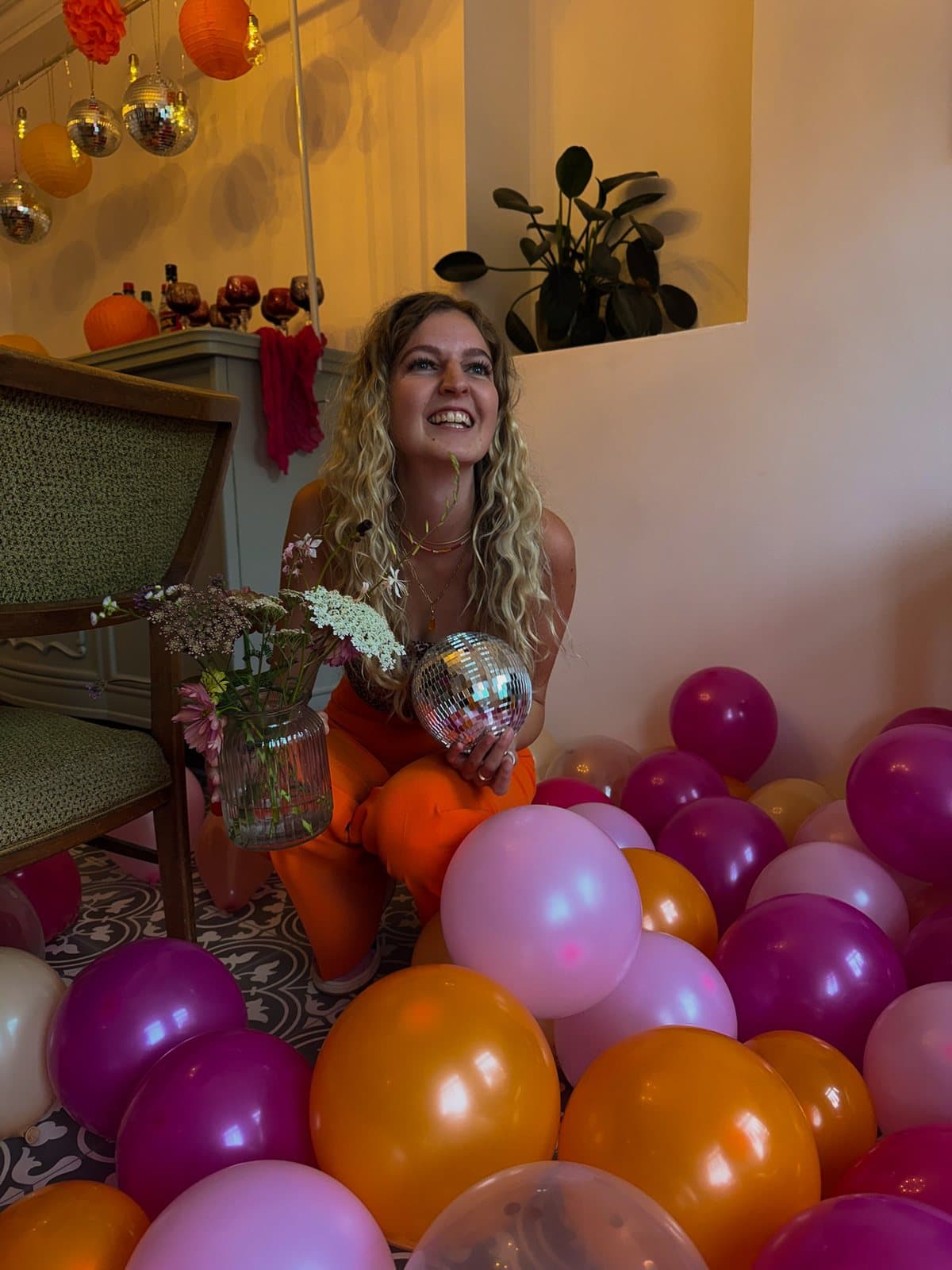 Lachende Frau mit Diskokugel und Blumenvase zwischen vielen pinken und orangefarbenen Luftballons