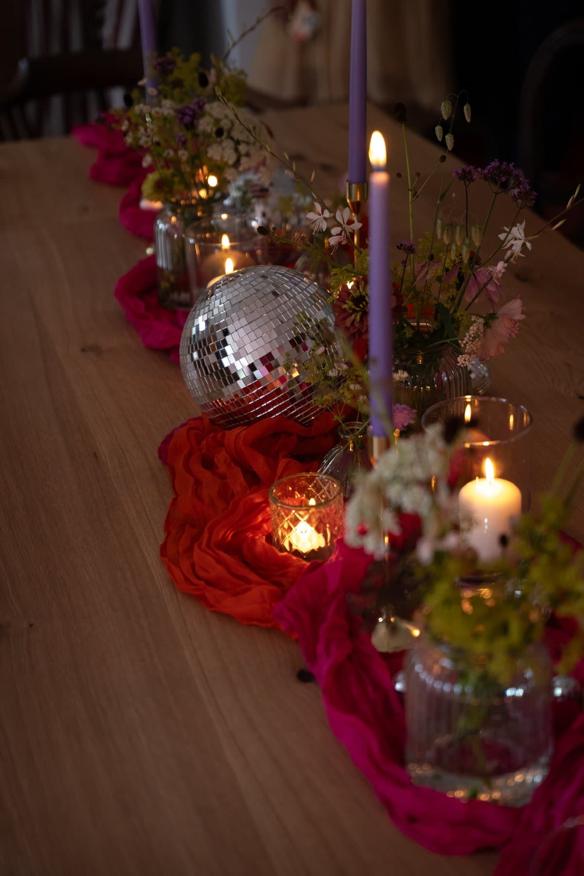 Holztafel mit rotem und pinkem Tischläufer, violetten Kerzen, Wiesenblumen und Diskokugel