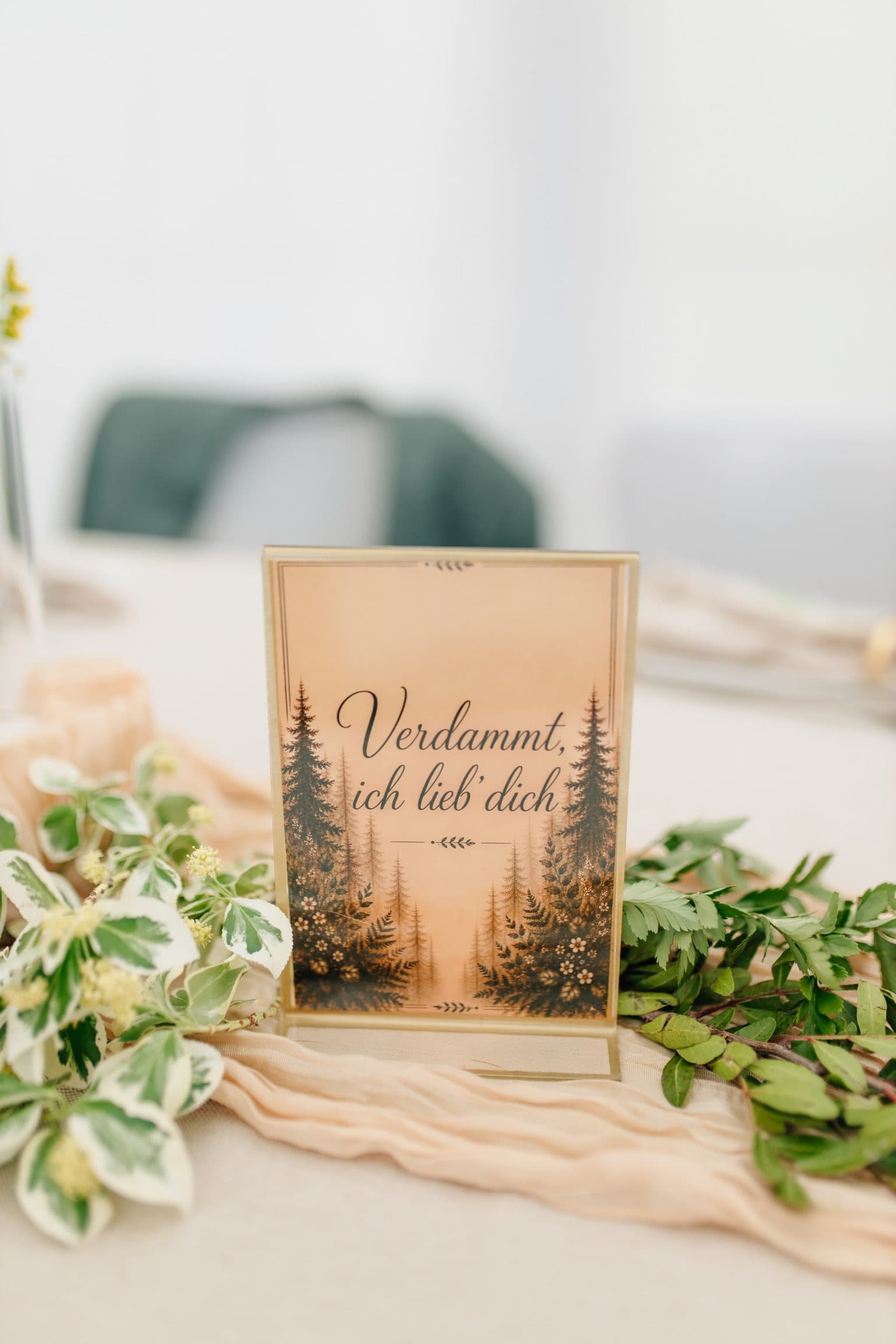 Tischschild im Goldrahmen mit Tannenwald-Motiv und der Aufschrift „Verdammt, ich lieb' dich“ zwischen Efeu und Tischläufer